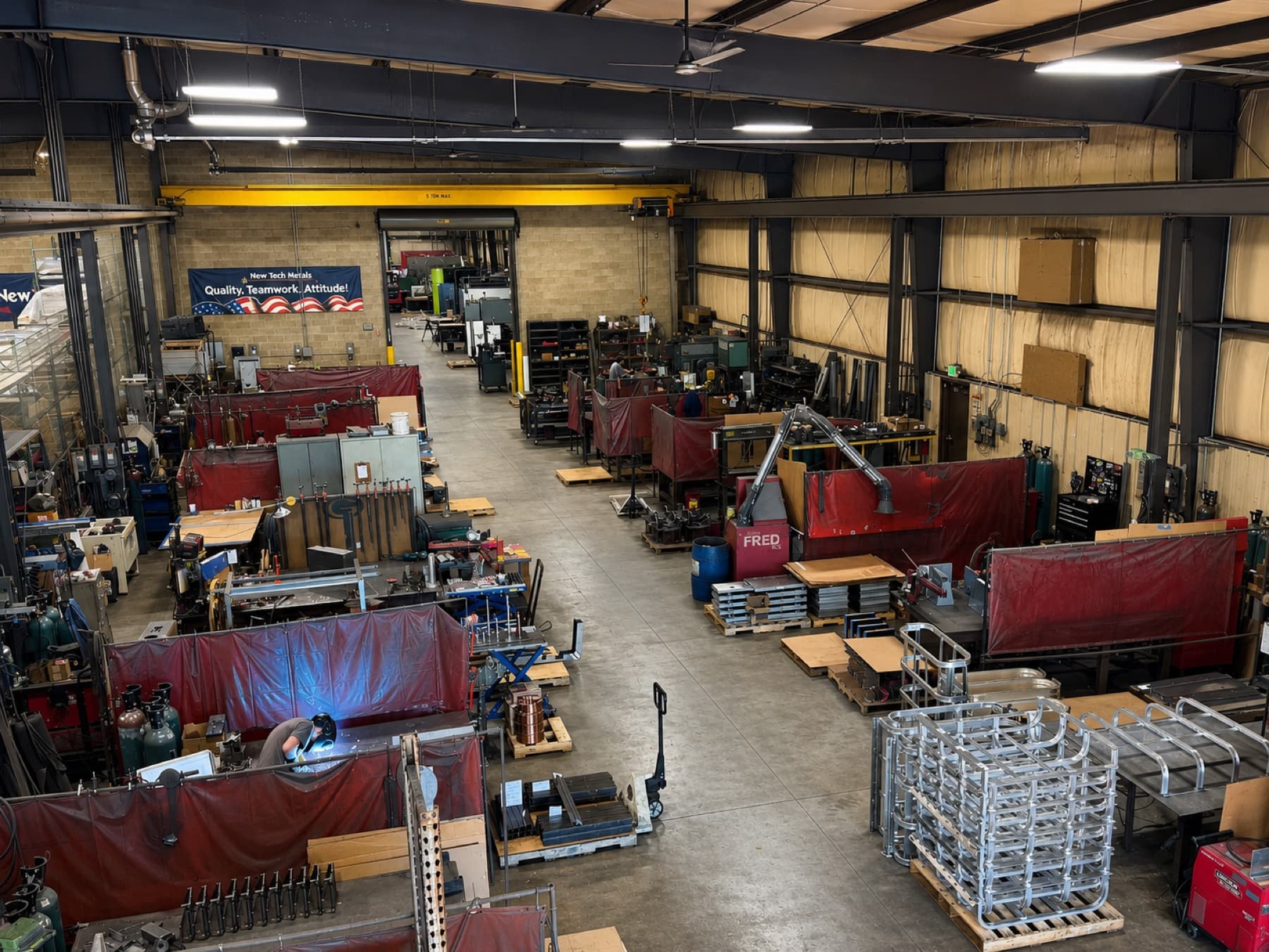 Overhead view of the New Tech Metals welding area with curtained weld stations, fume extraction, and a welder at work beneath the 5-ton overhead crane